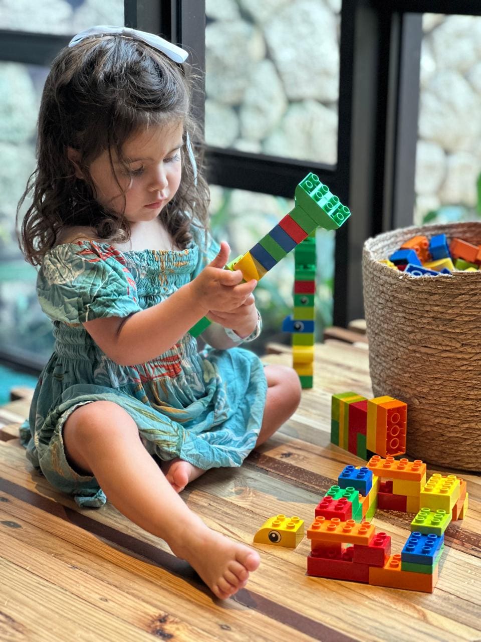 Young child in a green dress concentrating on building a creature with colourful Duplo blocks